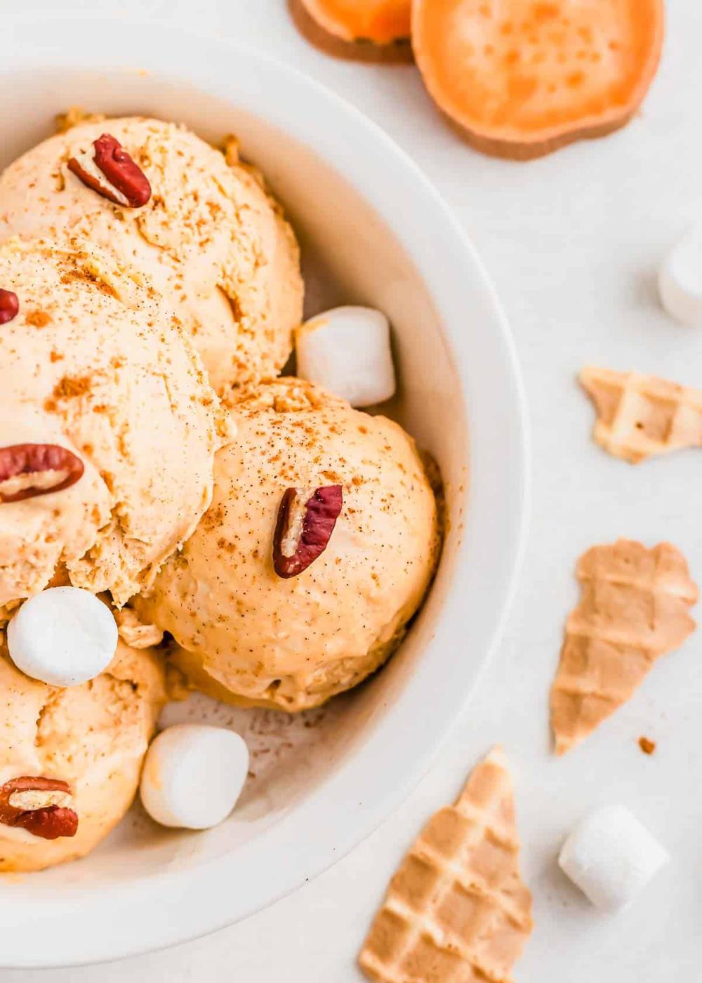 A bowl of sweet potato ice cream garnished with pecans and marshmallows. Waffle cone pieces and additional marshmallows are scattered nearby on a white surface.