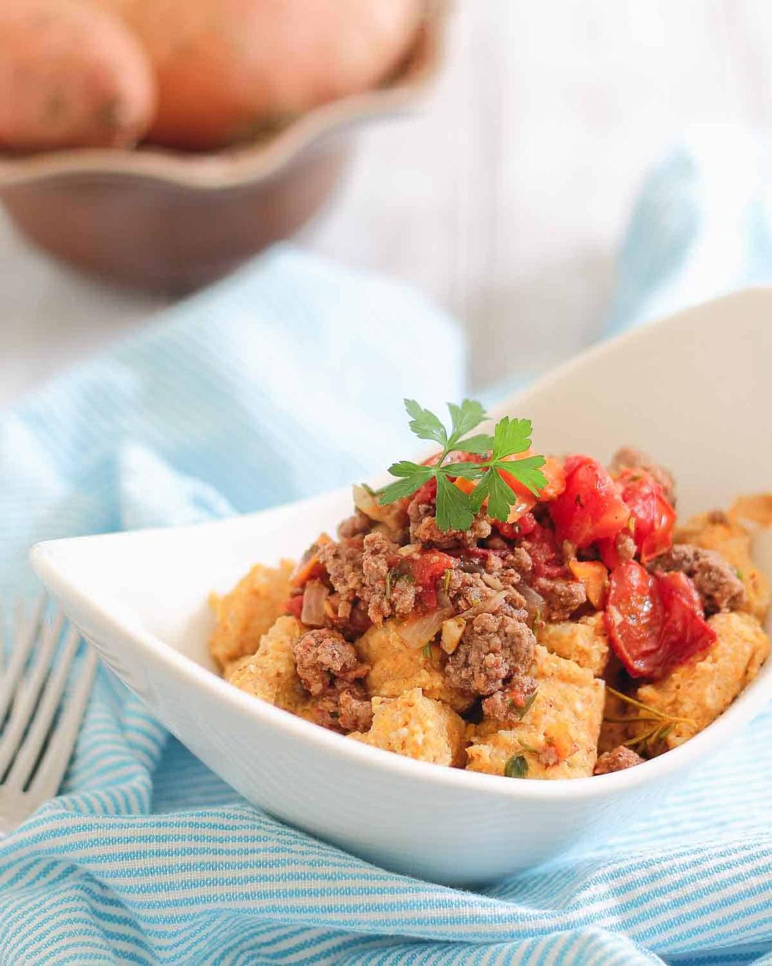 A white bowl filled with sweet potato gnocchi topped with cooked ground meat and cherry tomatoes, garnished with parsley. A blue striped cloth and a fork are next to the bowl.