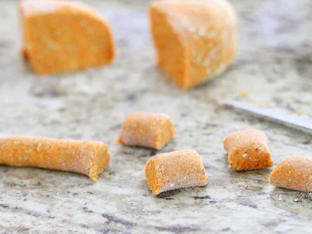 Close-up of raw gnocchi dough pieces being cut on a marble countertop, with some rolled dough and a knife visible in the background.