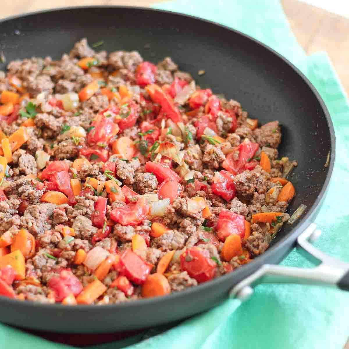 A skillet filled with cooked ground meat, diced tomatoes, carrots, and onions on a green napkin.