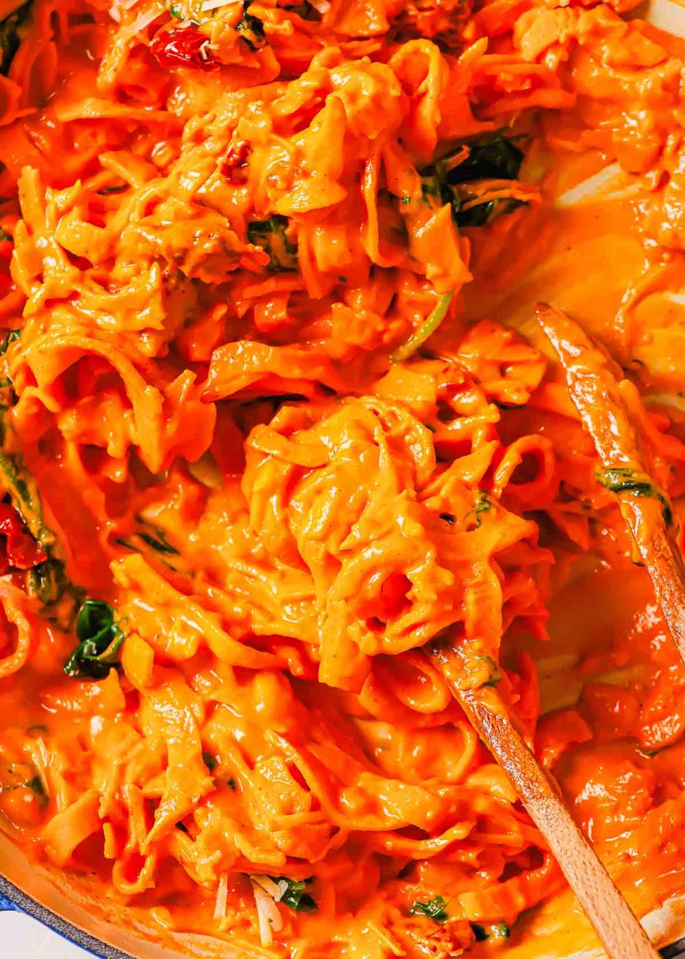 Close-up of sweet potato noodles mixed with a creamy sauce, spinach and sun-dried tomatoes, with two wooden spoons for stirring.