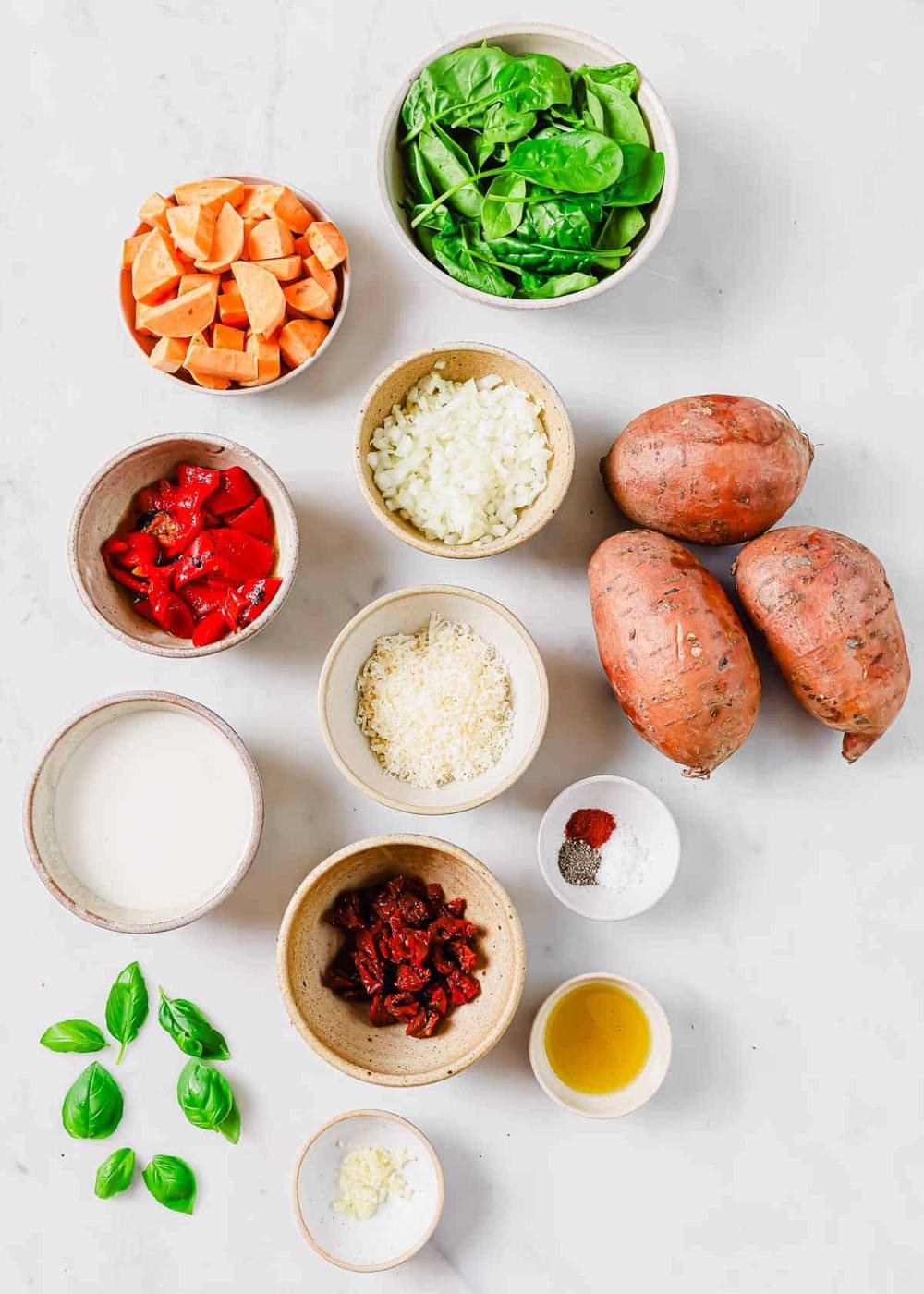 An assortment of ingredients including spinach, sweet potatoes, chopped onions, red peppers, milk, grated cheese, sun-dried tomatoes, basil leaves, peeled garlic, and seasonings arranged on a white surface.