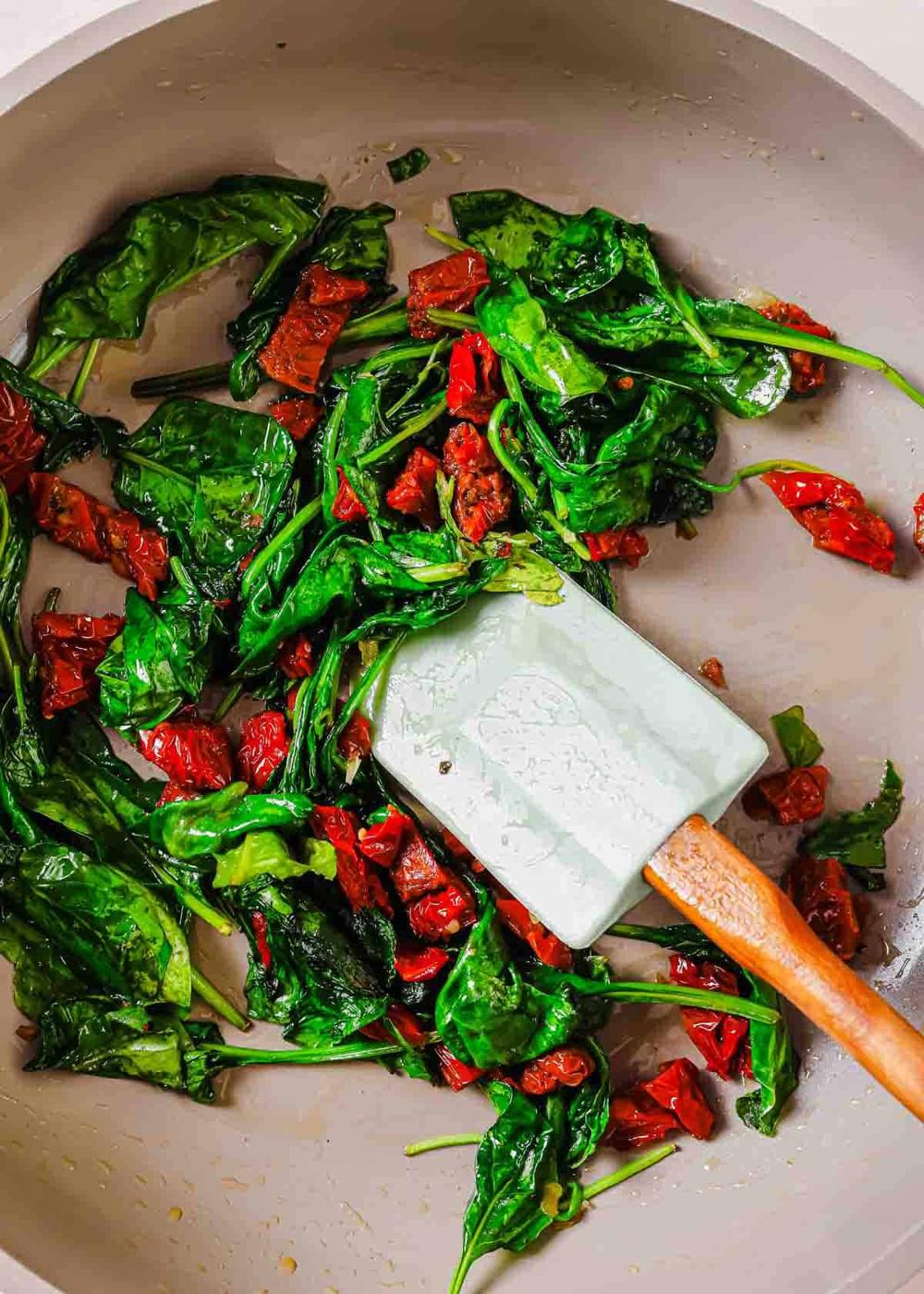 A frying pan contains wilted spinach and pieces of sun-dried tomatoes being stirred with a white spatula.