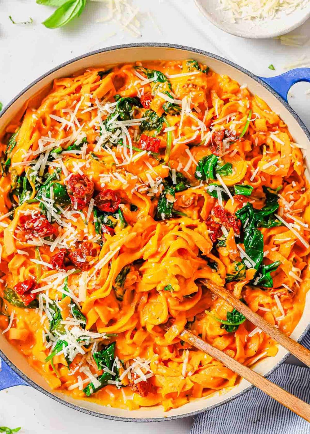 A skillet of cooked sweet potato pasta in a creamy sauce with spinach, sun-dried tomatoes, and topped with grated Parmesan cheese. Wooden tongs are in the pasta.