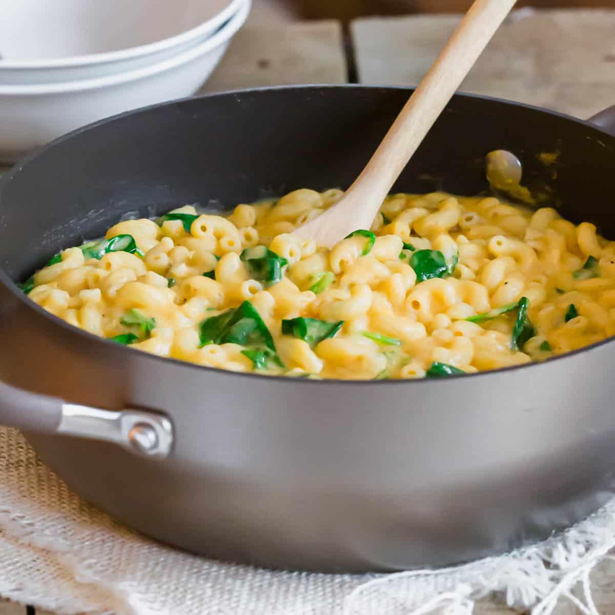 A pot of creamy sweet potato macaroni and cheese with spinach, stirred by a wooden spoon, placed on a textured cloth. In the background, there are two stacked white bowls on a wooden surface.