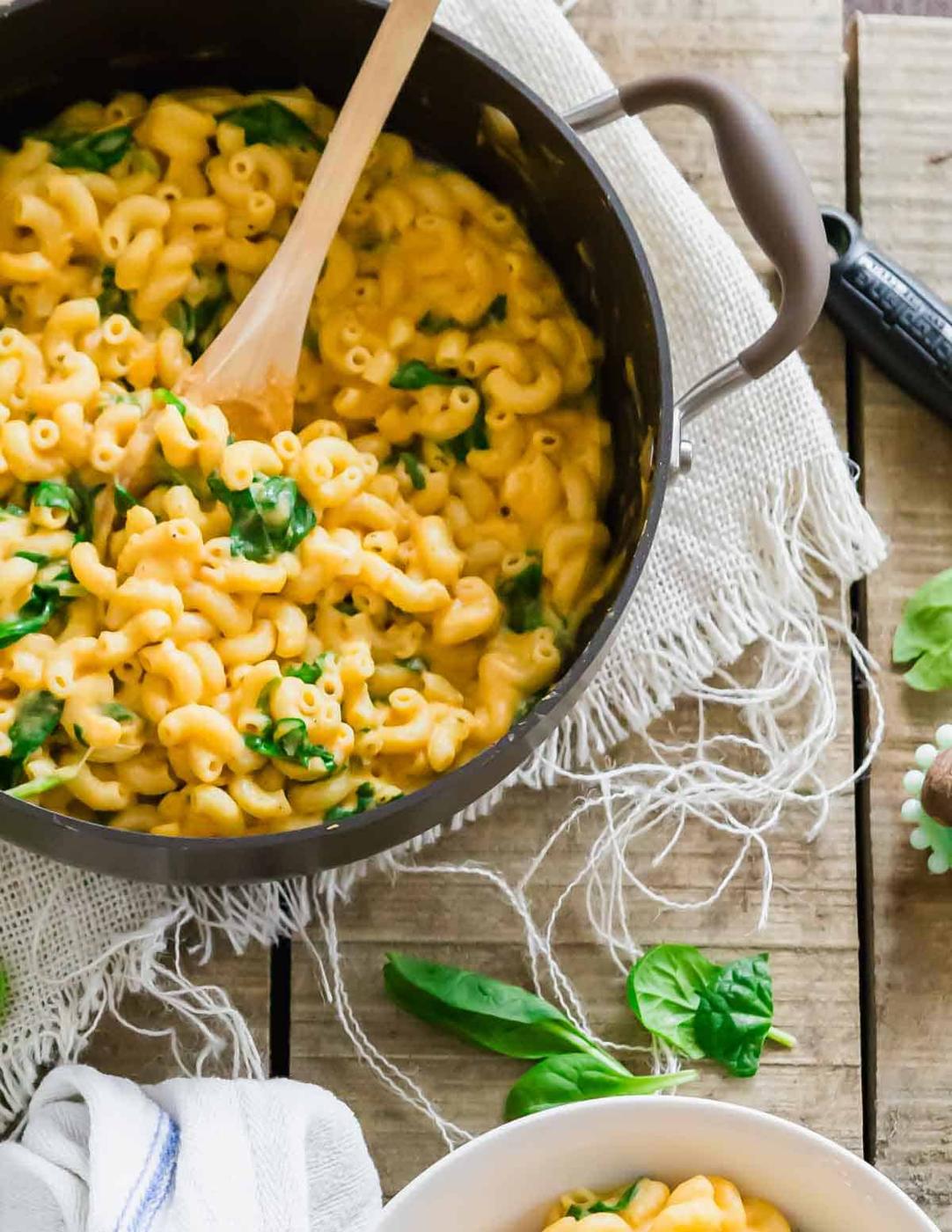 A pot of creamy sweet potato mac and cheese with spinach on a rustic wooden table. A wooden spoon is in the pot, and a white cloth is nearby. A few fresh spinach leaves are scattered around the table.