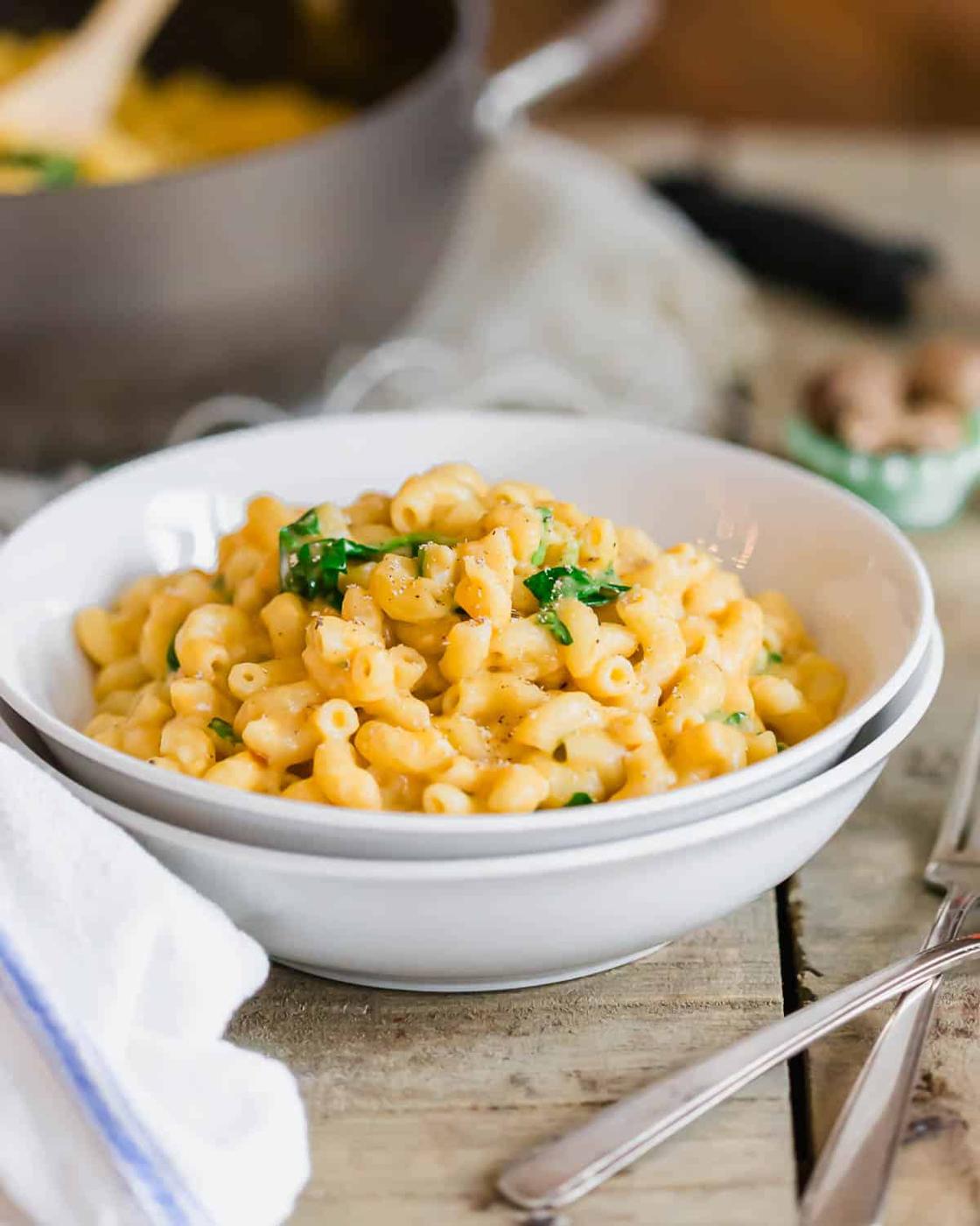 A white bowl filled with creamy macaroni and cheese and wilted spinach sits on a rustic wooden table. Nearby are silverware and a white napkin. In the background, there's a hint of a cooking pot.