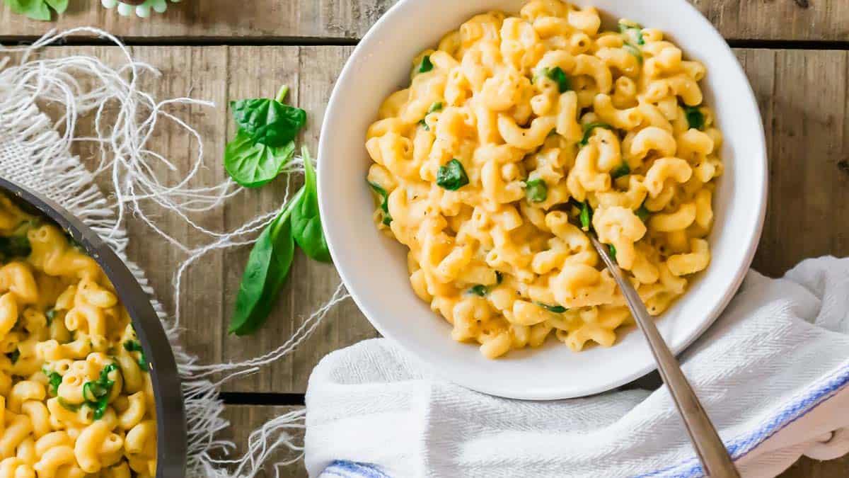 A bowl of macaroni and cheese made with creamy sweet potato sauce with baby spinach on a wooden table. A fork rests in the bowl, and a striped cloth is nearby. A pan with more pasta and spinach leaves is visible beside the bowl.