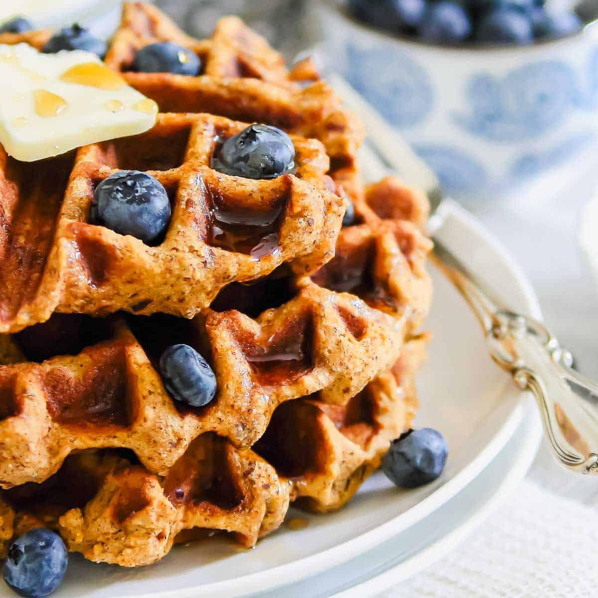 A stack of golden-brown waffles made with sweet potato topped with fresh blueberries and a pat of butter, drizzled with syrup, on a white plate. In the background, a bowl filled with more blueberries and a silver fork and knife are visible.
