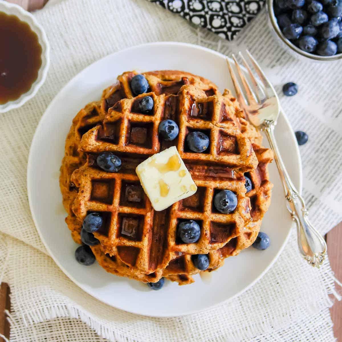 A plate of waffles topped with blueberries and a pat of butter, served with a silver fork and knife. A small bowl of syrup and a cup of blueberries are visible in the background. The setting includes a white, textured tablecloth.