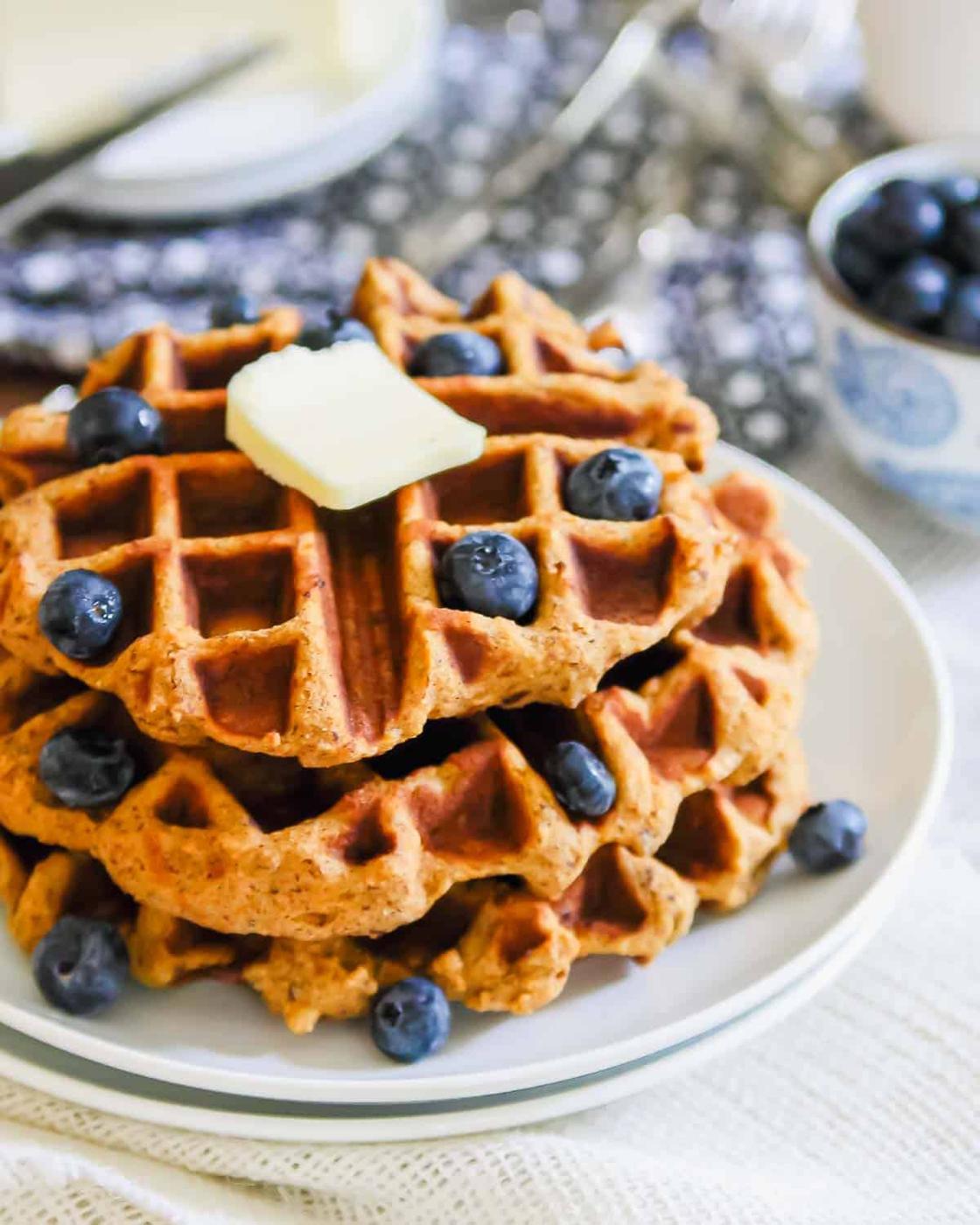 A stack of sweet potato waffles on a white plate, topped with a square of butter and scattered with fresh blueberries. A bowl of blueberries is in the background, sitting on a patterned tablecloth.