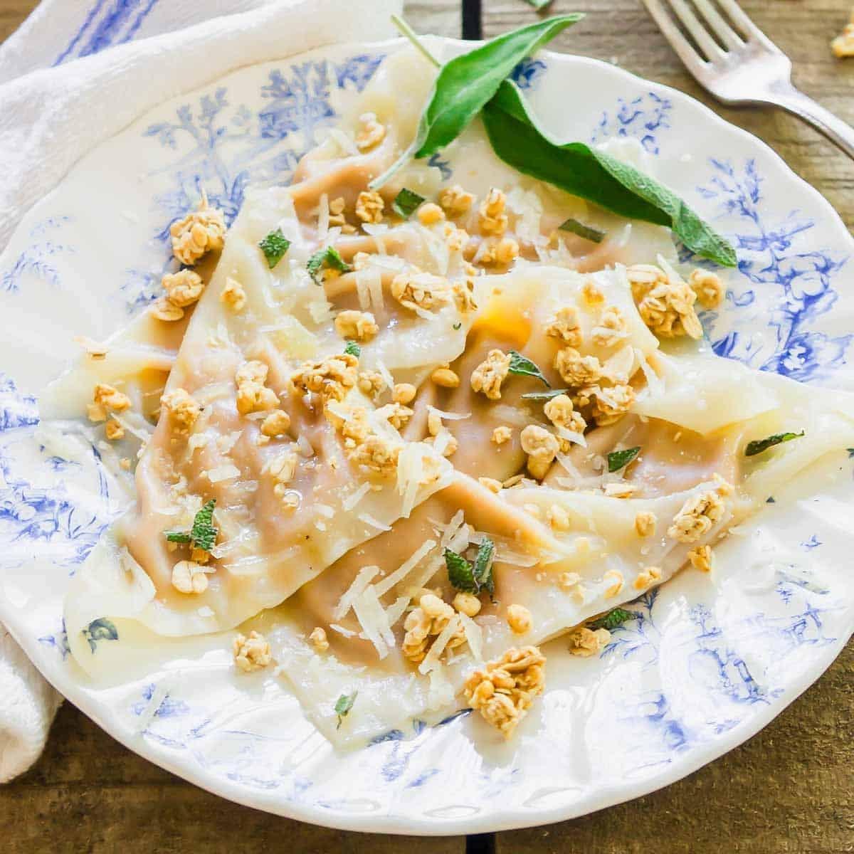 A plate with three folded ravioli topped with grated cheese, crispy sage, and crunchy bits, served on a blue and white patterned dish with a fork and cloth napkin nearby.