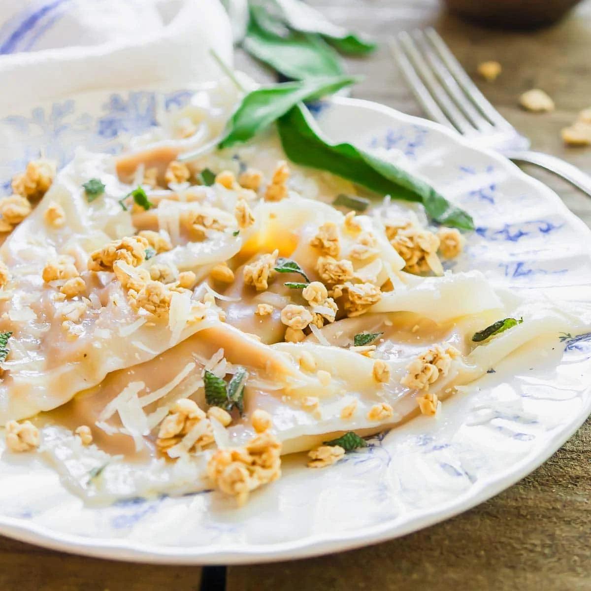 A close-up of homemade ravioli topped with grated cheese, fresh herbs, and crunchy granola, served on a decorative white and blue plate with a fork and napkin nearby.