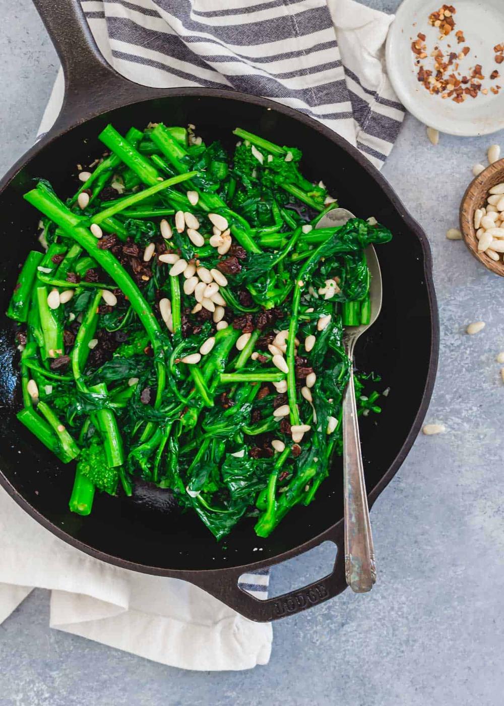 Broccoli rabe in a cast iron skillet with a spoon topped with pine nuts and raisins.