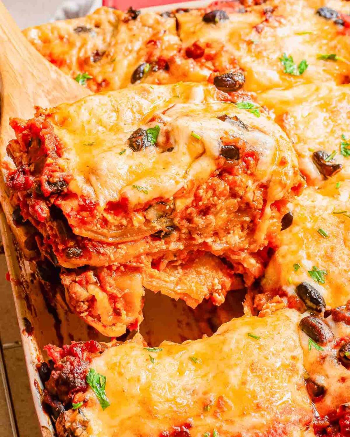 A serving of cheesy taco lasagna is being lifted out of a baking dish with a wooden spatula. The lasagna is topped with melted cheese, tomato sauce, and garnished with herbs.