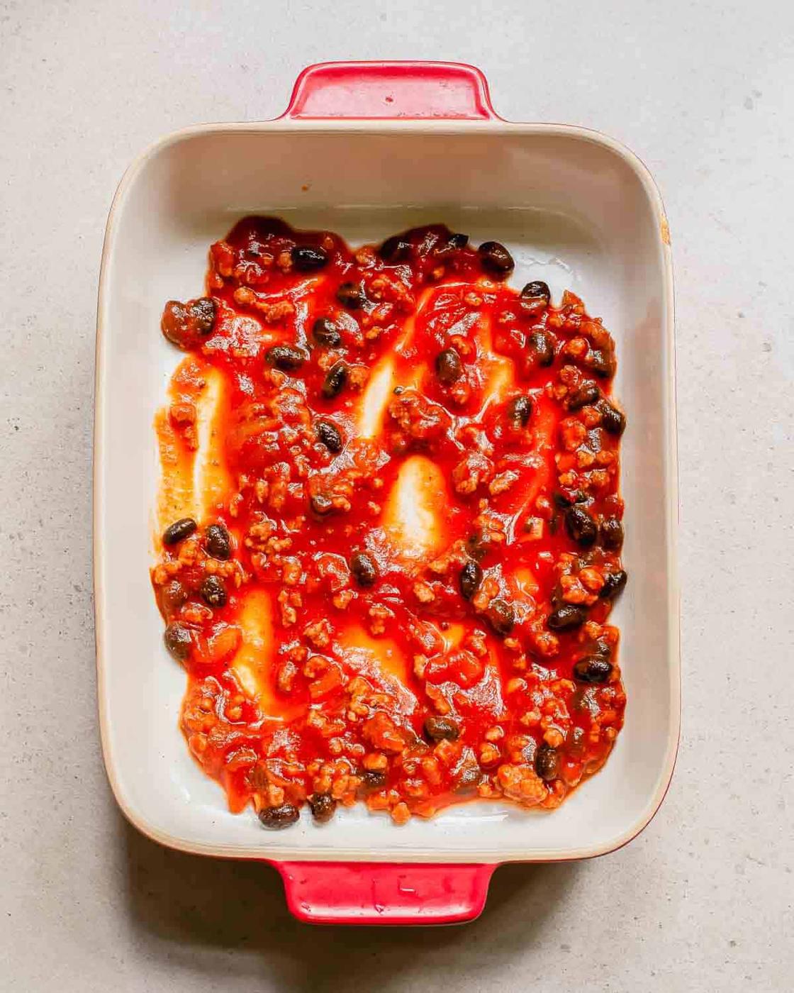A rectangular baking dish with red handles containing a mix of tomato sauce, black beans, and other visible ingredients spread unevenly.