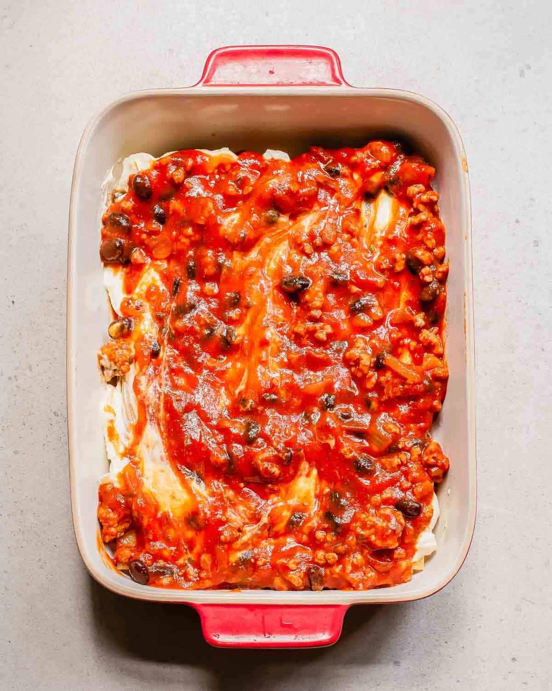 A rectangular baking dish with a partially assembled lasagna, showing layers of ricotta cheese and a tomato-based sauce with ground meat and black beans.
