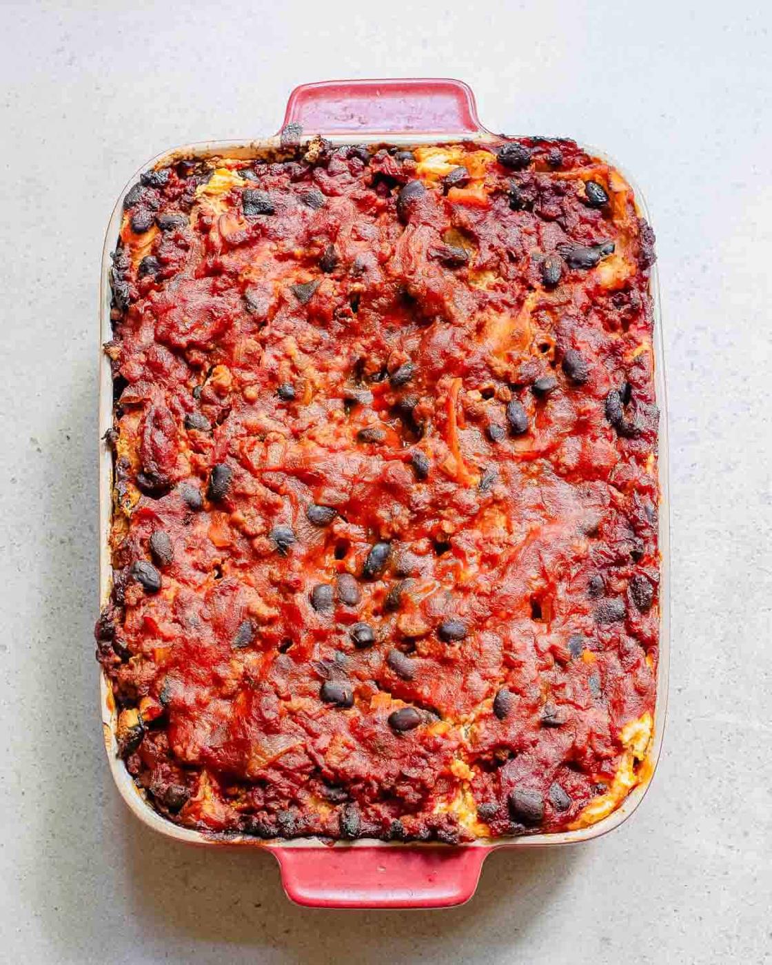 A baked lasagna in a rectangular red dish, topped with melted cheese and a layer of tomato sauce, on a light-colored surface.