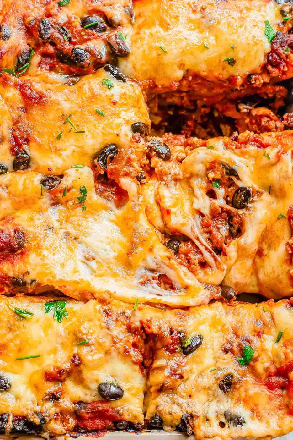Close-up of a cheesy lasagna with visible layers of pasta, ground meat, tomato sauce, black beans, and melted cheese, garnished with a sprinkle of fresh parsley.