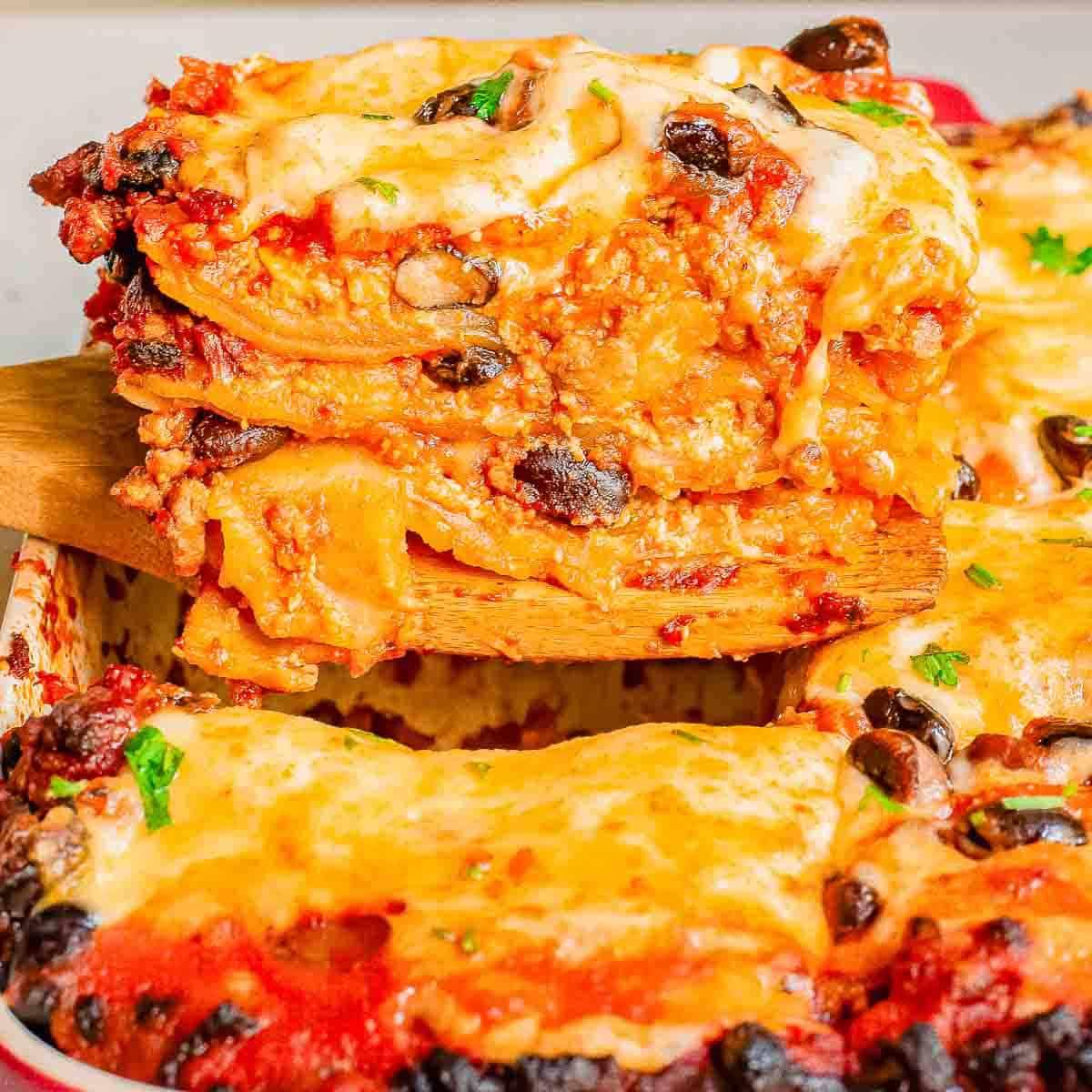 A close-up of a slice of Mexican lasagna being lifted from a casserole, featuring layers of pasta, tomato sauce, cheese, and vegetables.