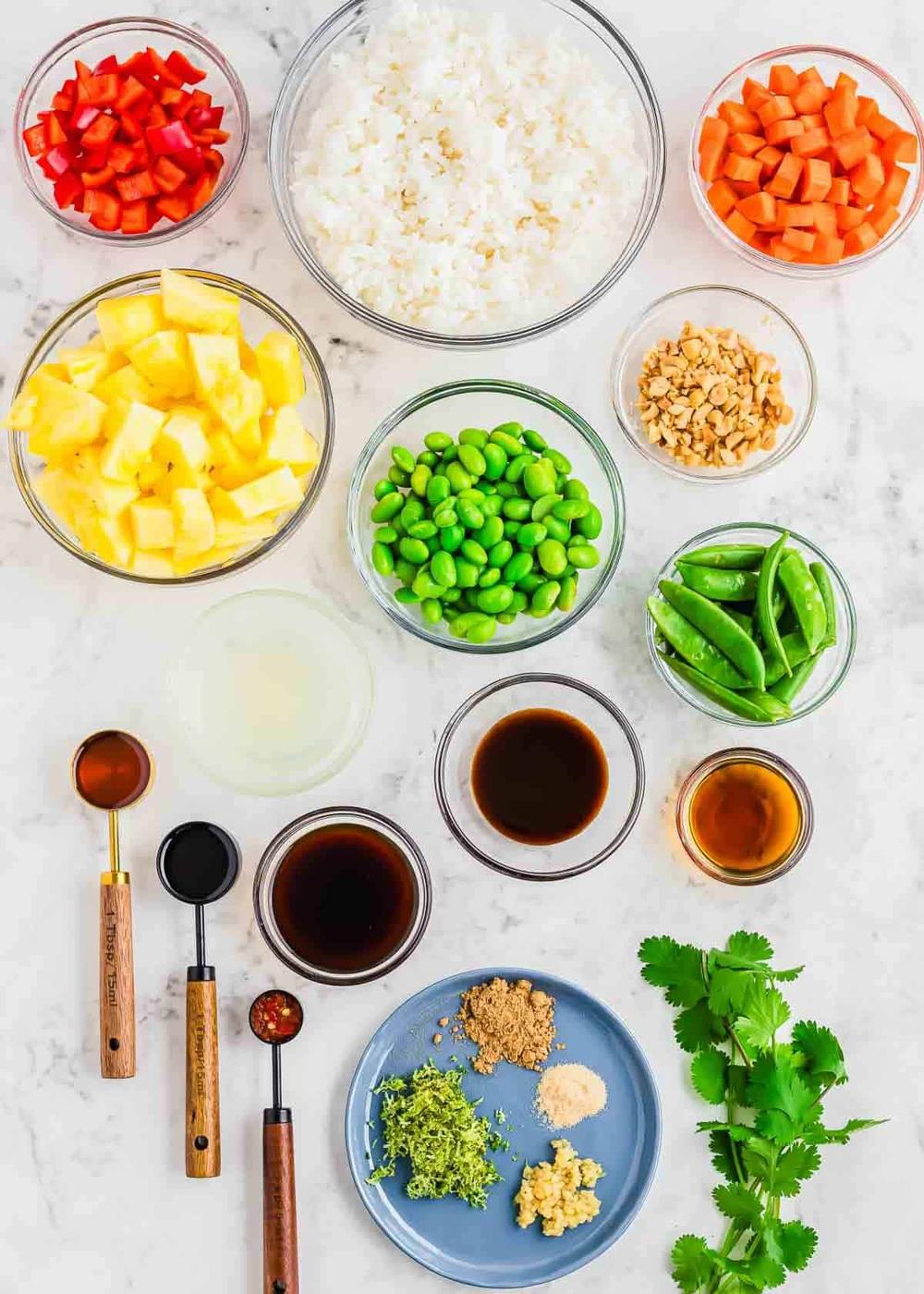Top view of assorted ingredients in bowls for a colorful fried rice dish on a white marble surface. Includes diced red peppers, rice, pineapple, carrots, edamame, peanuts, snap peas, lime juice, and various sauces and spices, with fresh cilantro on the side.