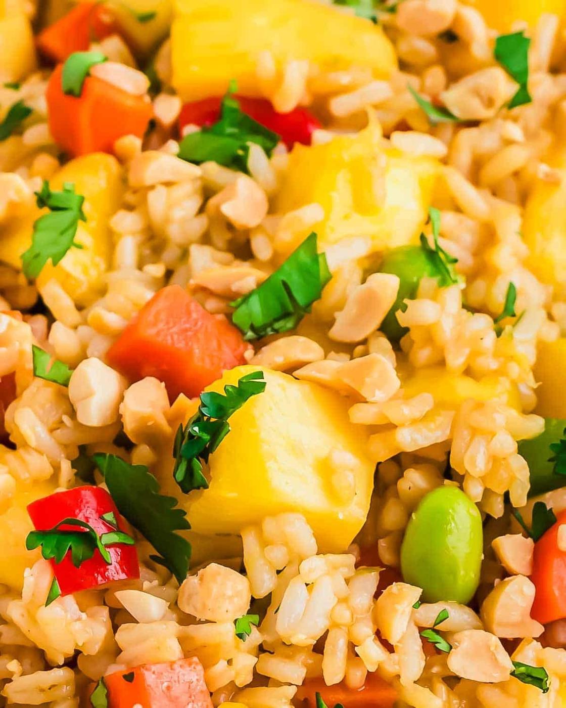 A vibrant close-up of pineapple fried rice dish featuring red bell peppers, carrots, edamame, and garnished with cilantro and crushed peanuts. The mix of ingredients creates a textured and appetizing appearance.