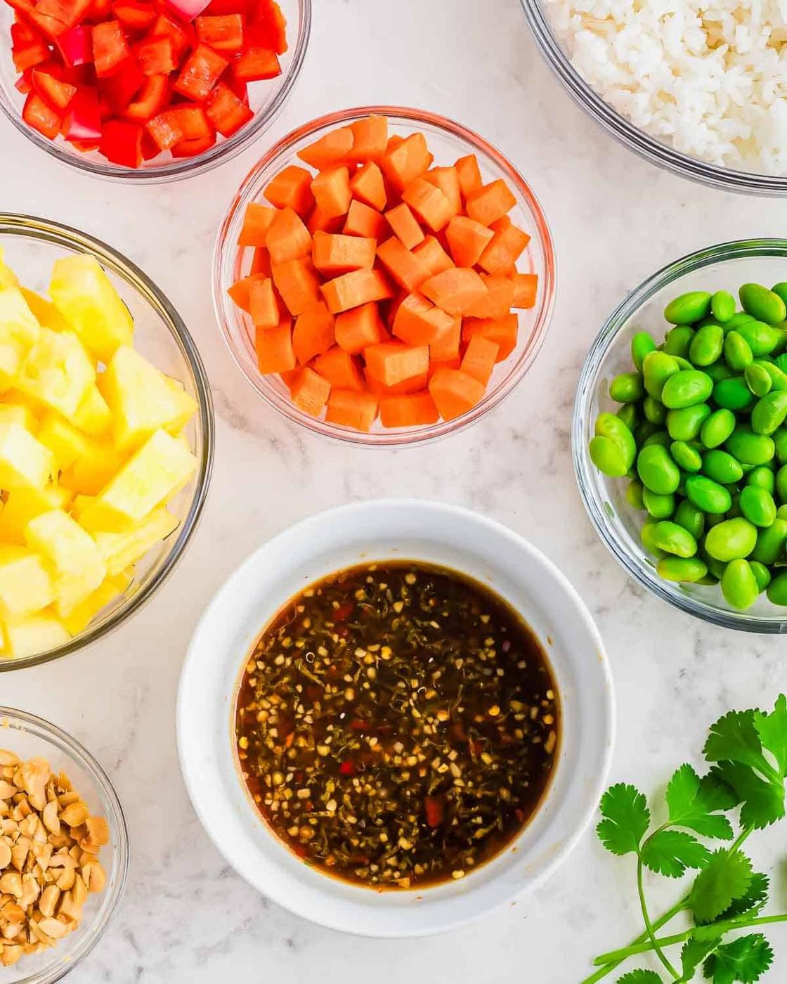 An array of ingredients in bowls: diced red bell peppers, cubed carrots, pineapple chunks, edamame beans, white rice, and crushed peanuts. In the center, a bowl of soy-based sauce with sesame seeds. Fresh cilantro sprigs are nearby. Overhead view.
