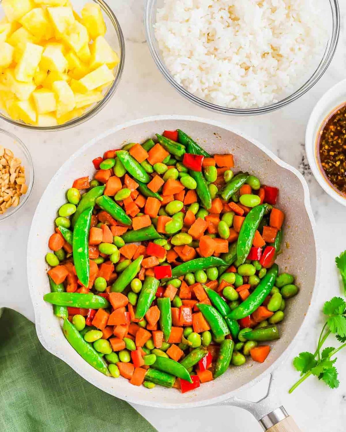 A colorful skillet with snap peas, edamame, carrots, and red bell peppers in a white skillet. Surrounding bowls contain diced pineapple, rice, crushed peanuts, and a brown sauce. A green napkin and cilantro garnish complete the setting.