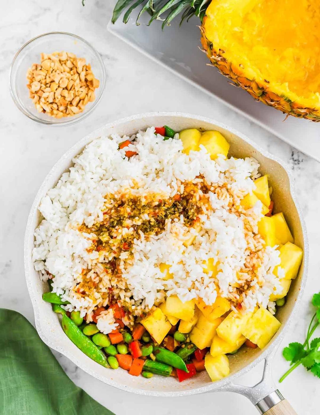 A skillet filled with white rice, pineapple chunks, and mixed vegetables, topped with sauce. A small bowl of chopped nuts is nearby. A halved pineapple with its flesh removed is in the background.