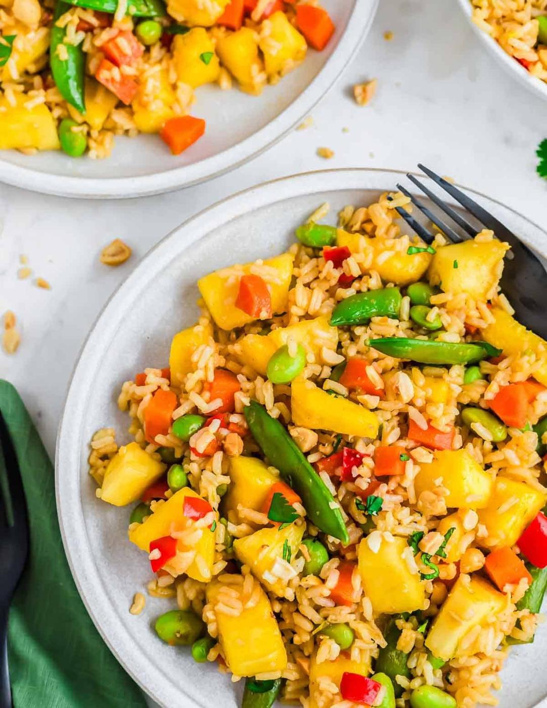 A plate of colorful pineapple fried rice with green beans, red bell peppers, edamame, and rice is garnished with chopped herbs. Another plate with the same dish is partially visible in the background, along with a black fork.