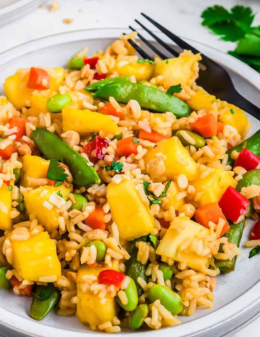 Pineapple fried rice recipe on a white plate featuring green sugar snap peas, edamame, red bell pepper pieces, and topped with chopped peanuts and fresh cilantro. A black fork is placed on the plate.