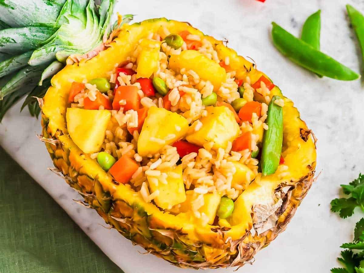 A pineapple half used as a bowl, filled with fried rice, pineapple chunks, peas, carrots, and green beans. The dish is placed on a white surface with sugar snap peas and parsley nearby.