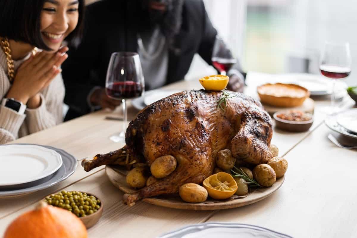 Joyful people are gathered around a table enjoying a traditional Thanksgiving feast with roasted turkey garnished with lemons and potatoes. Glasses of red wine accompany the meal, while dishes of peas, pie, and even some Thanksgiving feast alternatives like a pumpkin dish are also visible.