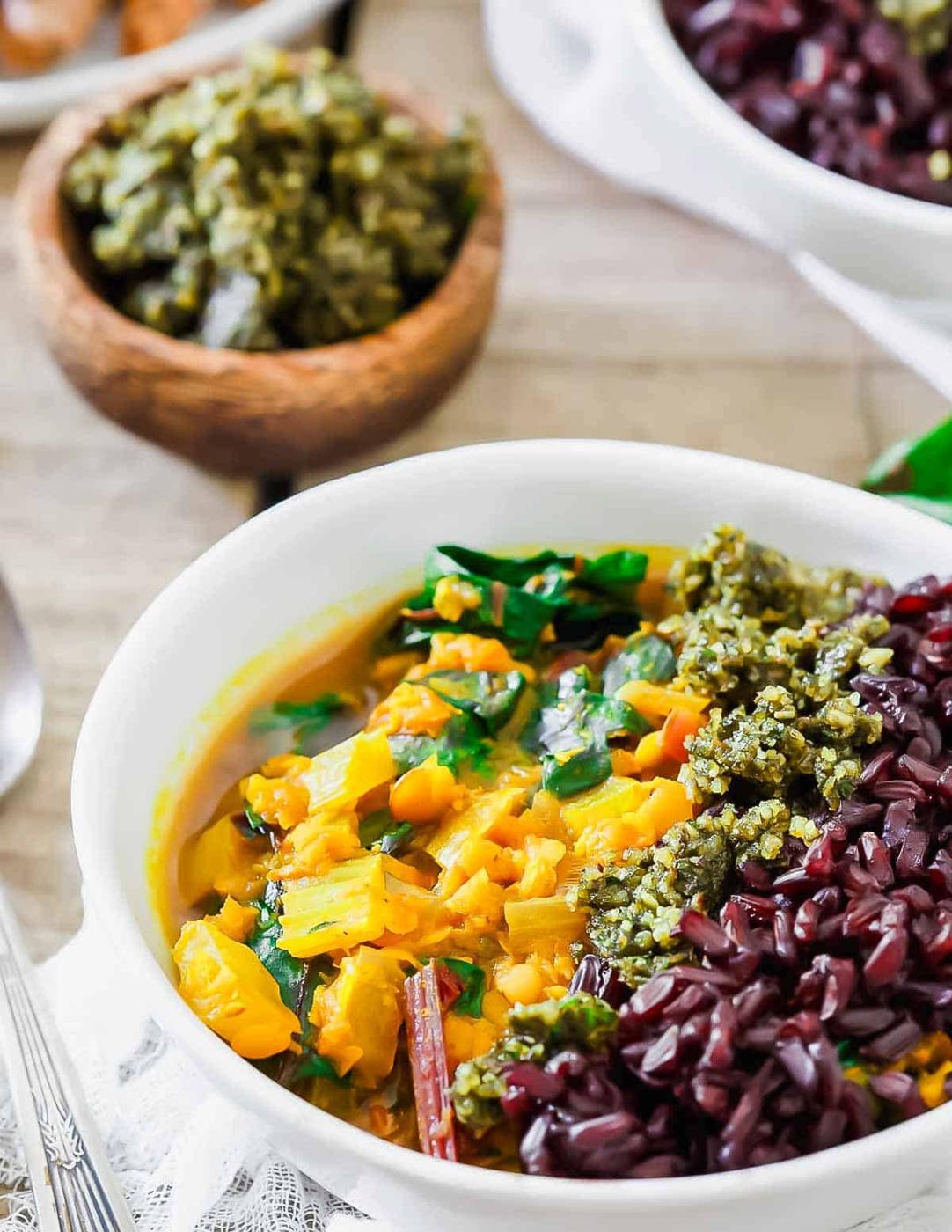 A vibrant lentil stew dish featuring purple rice and a green pesto-like topping in a white bowl. A wooden bowl filled with a similar green topping is in the background.