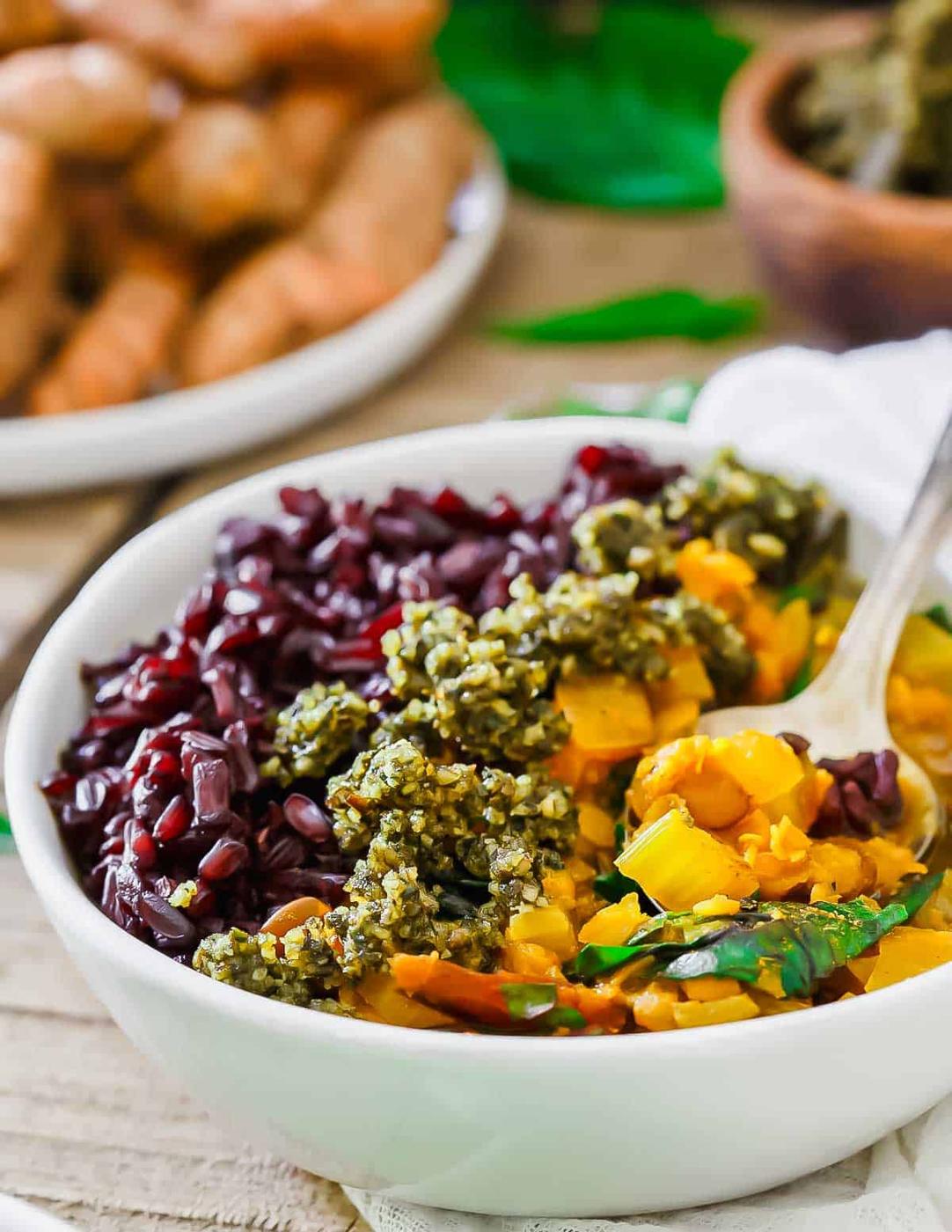 A bowl of black rice topped with lentil stew. In the background, a plate of fresh turmeric and a wooden bowl are partially visible.