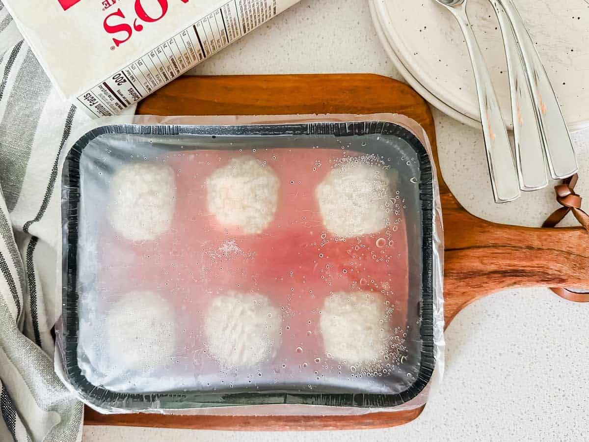 A frozen entree of six meatballs in sauce covered with plastic wrap sits on a wooden board next to a striped towel, a carton, a plate, and two metal forks.