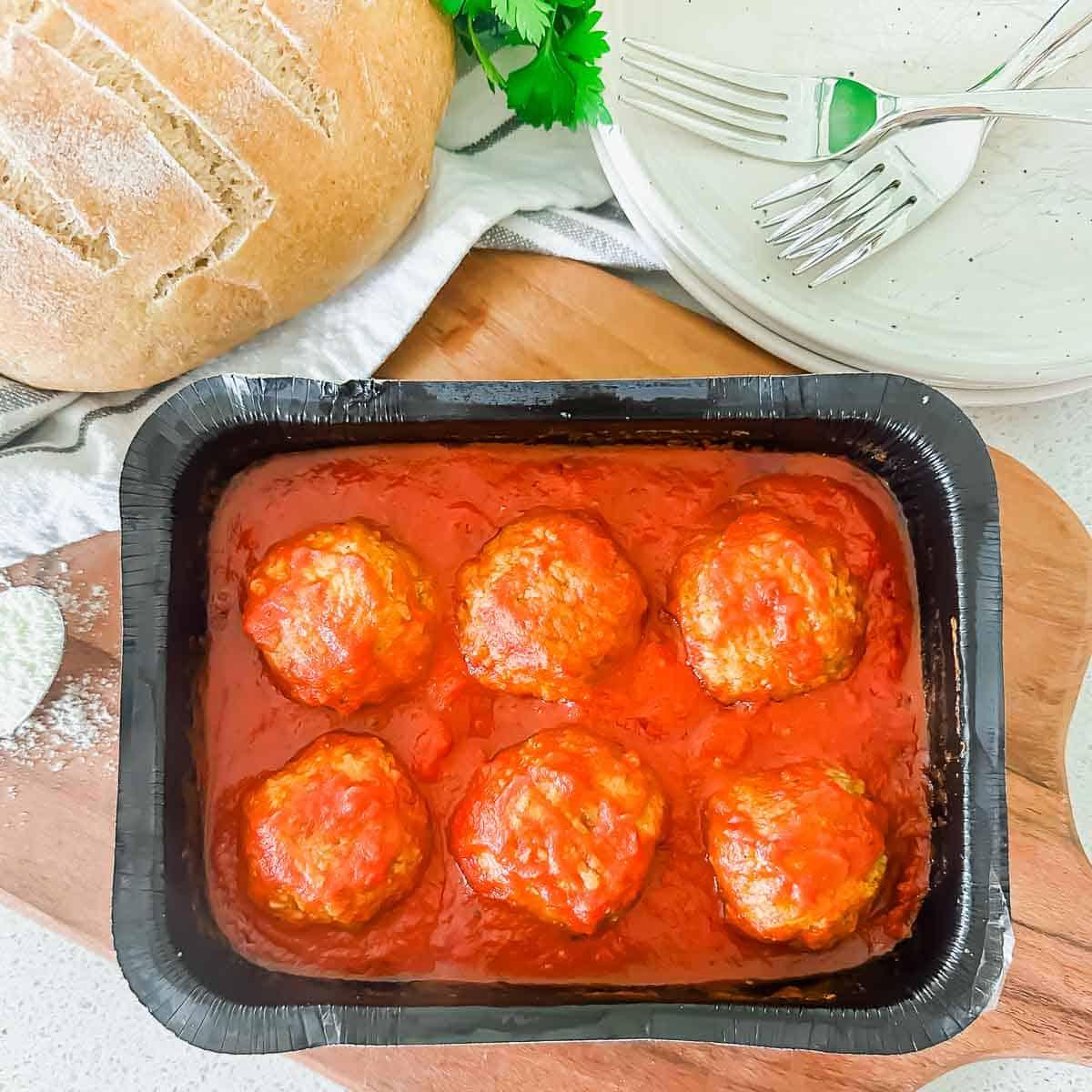 A black frozen dinner tray with six meatballs in tomato sauce sits on a wooden board, next to a loaf of bread, stacked white plates, and forks. Fresh herbs are partly visible in the background.