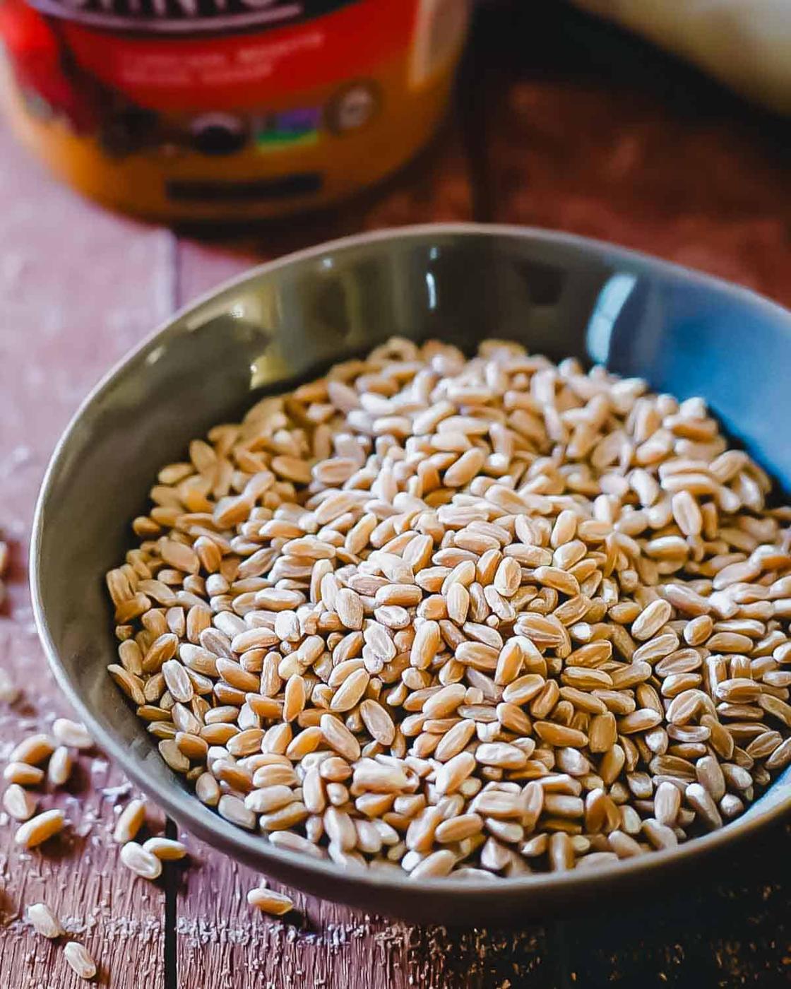 A gray bowl filled with farro on a wooden table. Some extra farro grains are scattered on the table around the bowl, and a blurred container is in the background.