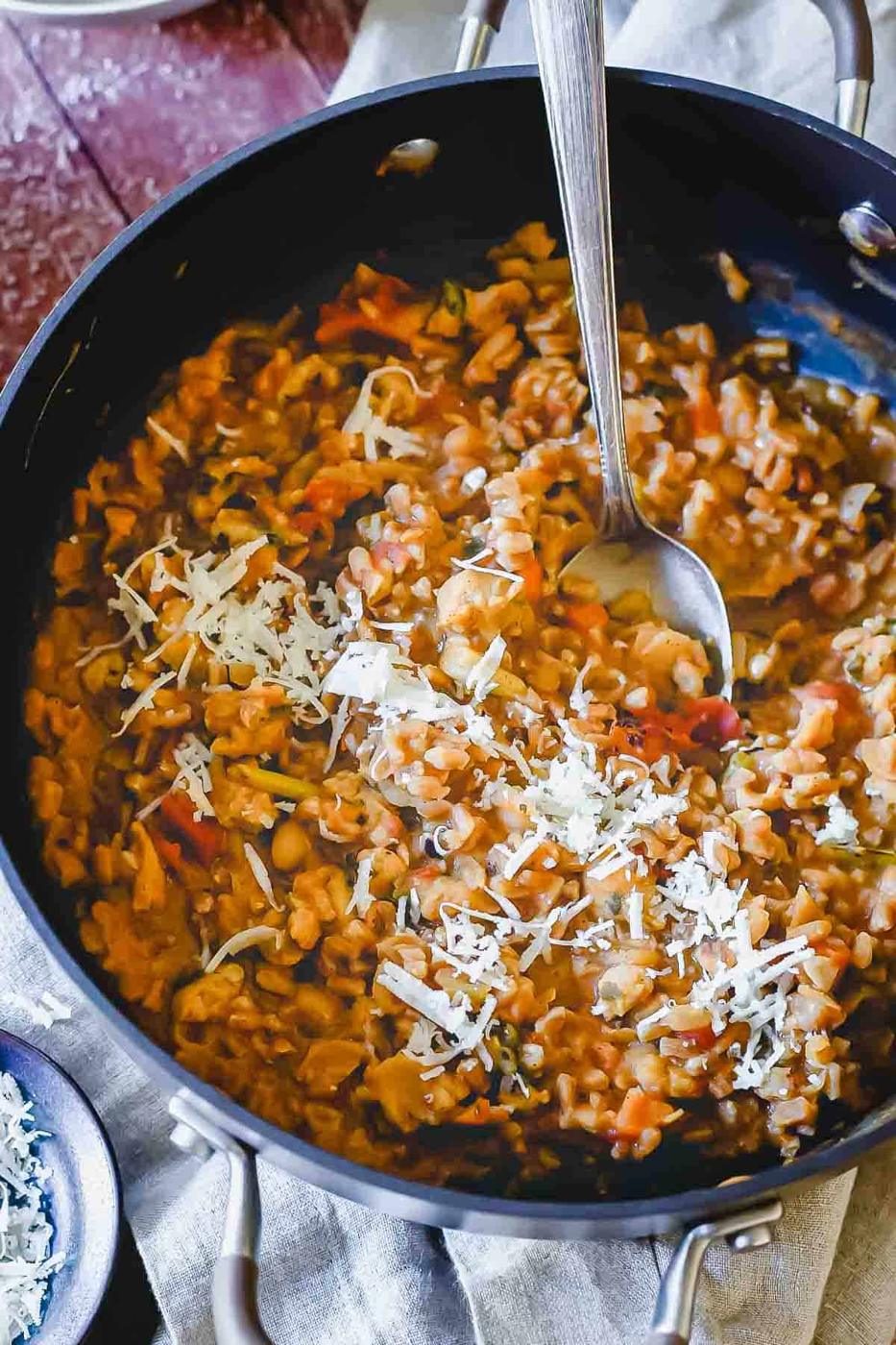 A pan filled with creamy farro risotto mixed with vegetables like carrots and spinach, topped with grated cheese. A spoon is placed in the pan, ready for serving.