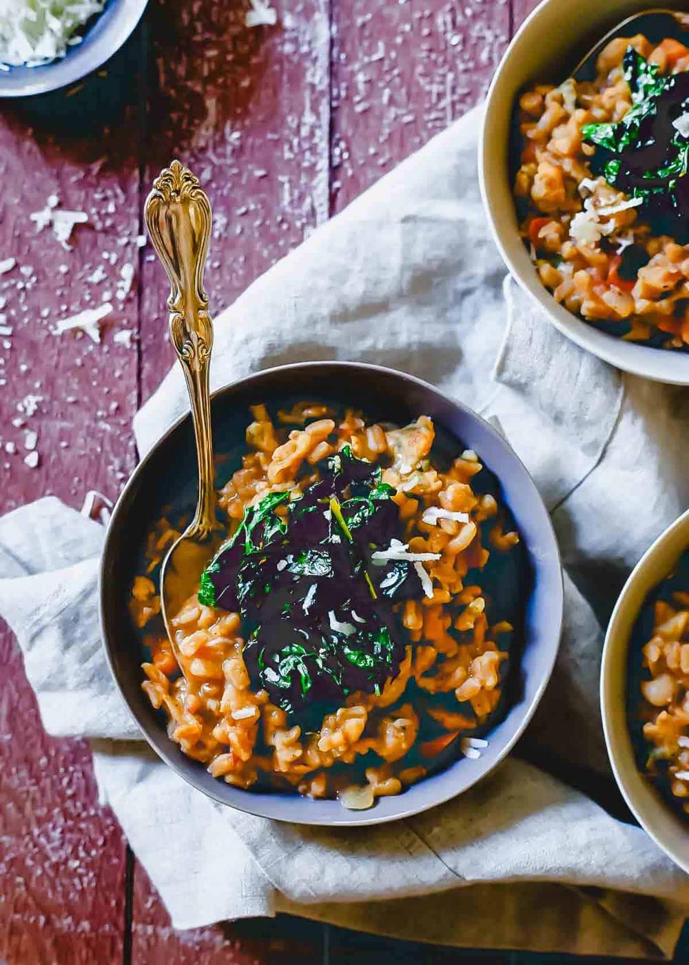 A bowl of farro risotto topped with dark leafy greens and shredded cheese, placed on a textured cloth. A gold spoon sits in the bowl. Another bowl and a small dish with cheese are nearby on a rustic wooden surface.