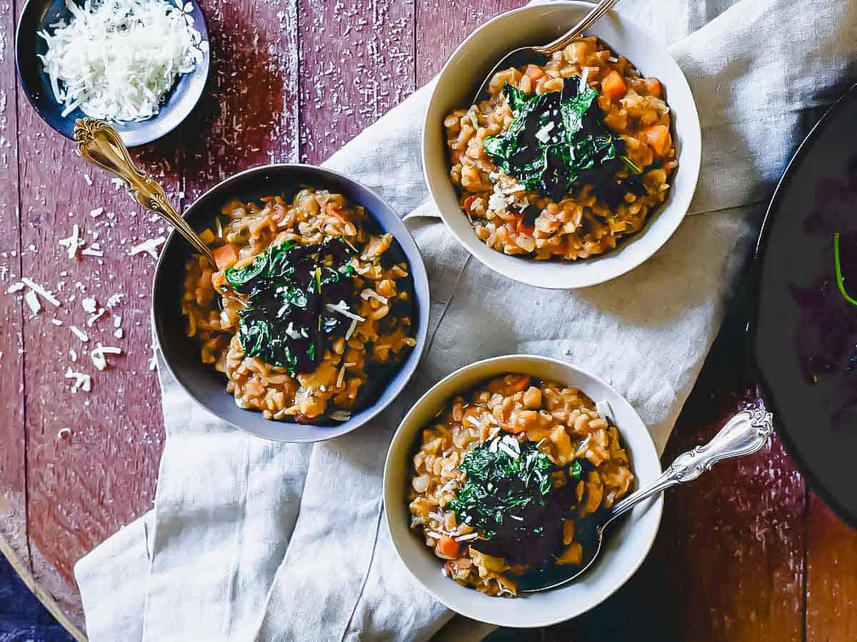 Three bowls of risotto garnished with greens are arranged on a rustic wooden table. A small dish of grated cheese is beside them. The setting has a cozy and inviting ambiance.
