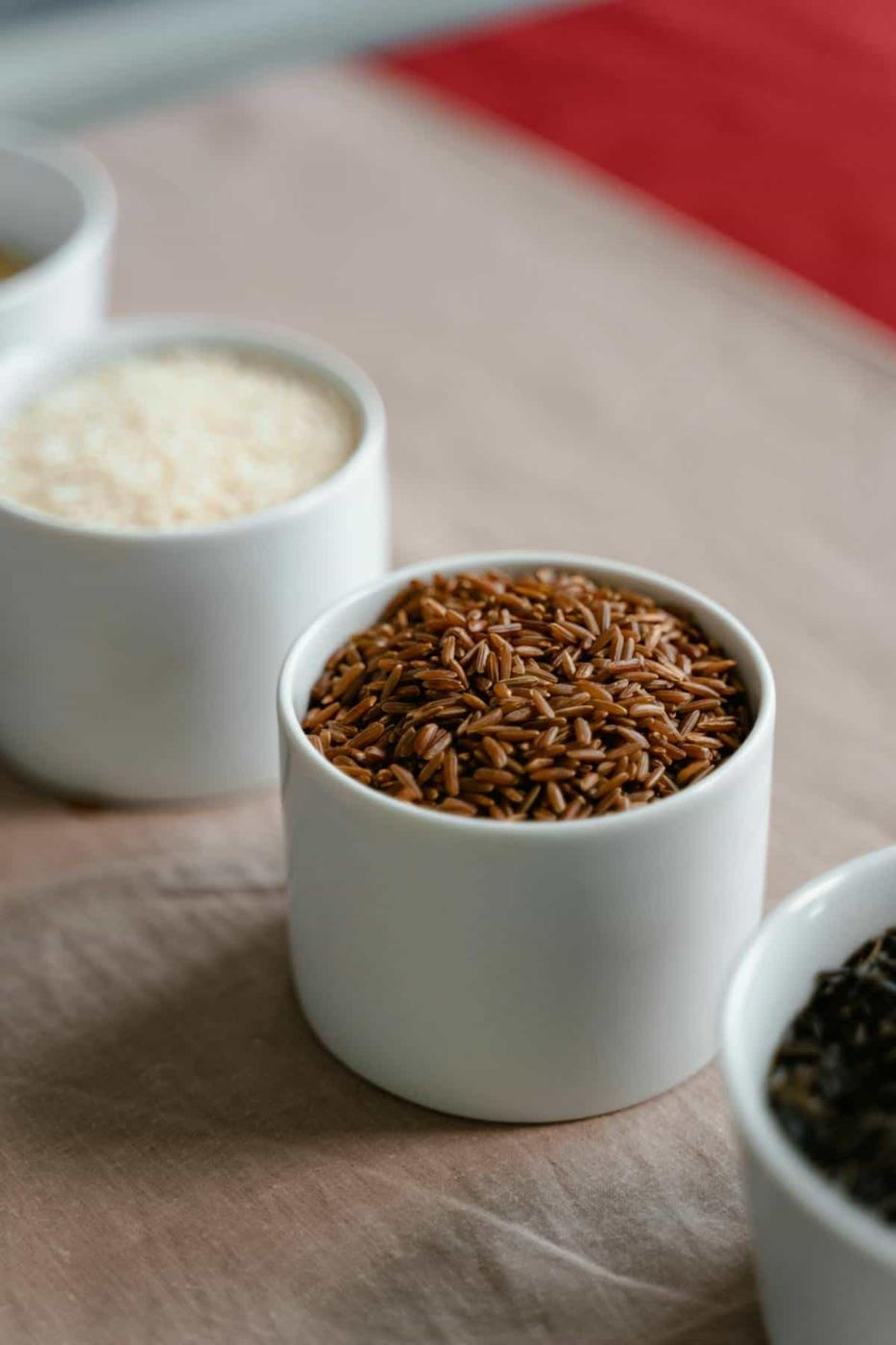 A close-up of a white bowl filled with brown rice on a table, with other bowls of various types of rice blurred in the background.