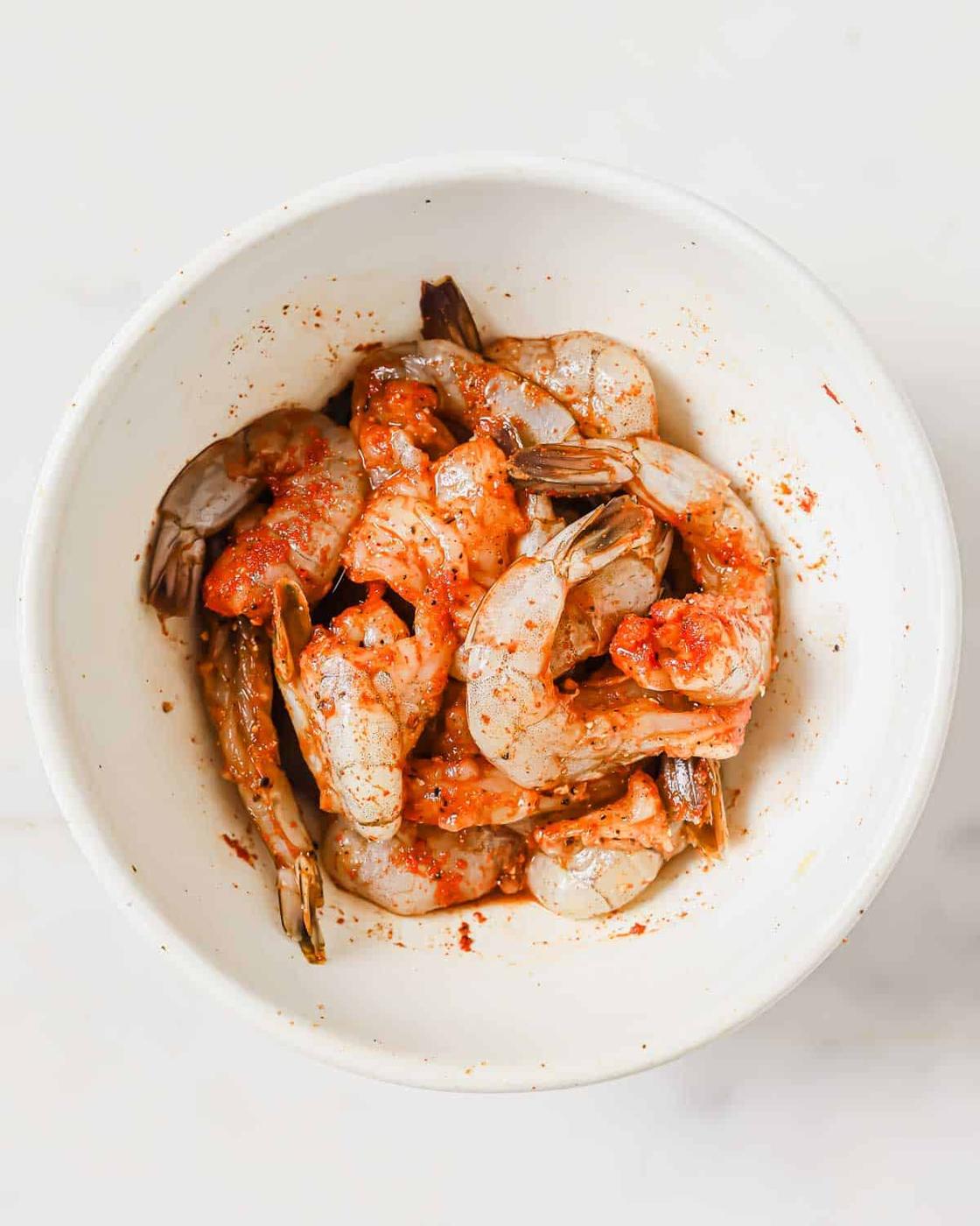 A white bowl filled with raw shrimp seasoned with spices.