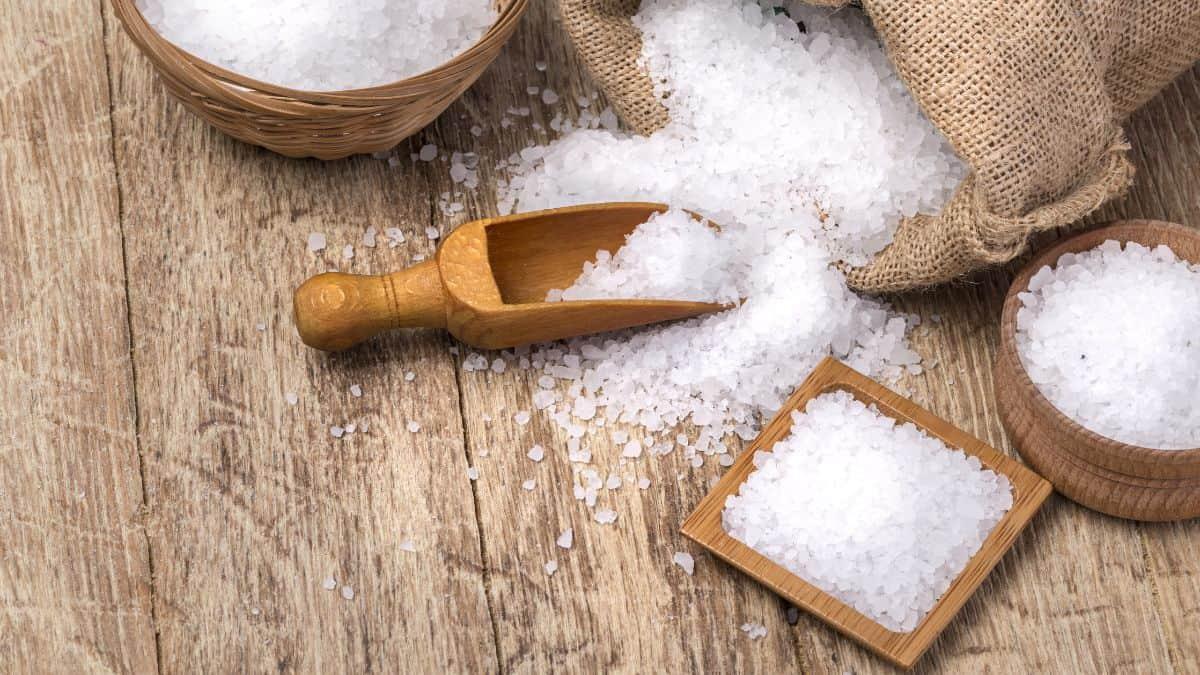 A wooden scoop and small bowls filled with coarse sea salt, arranged on a rustic wooden surface.