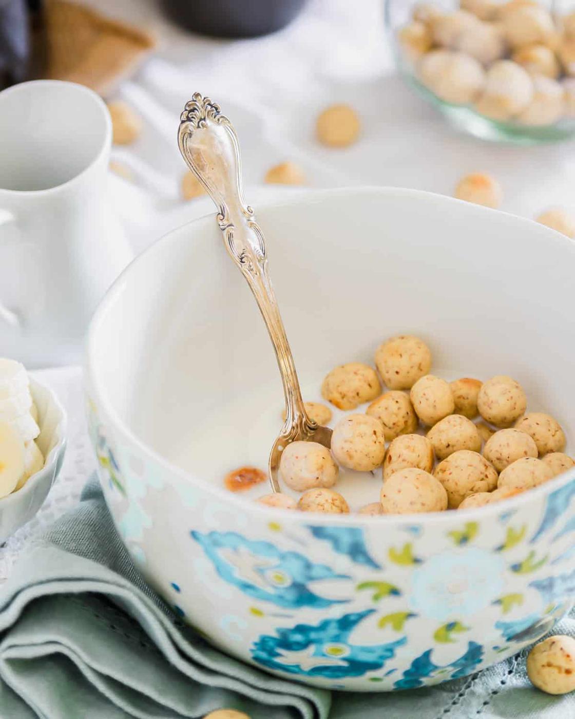 A large bowl with a floral design holds milk and homemade cereal puffs, with a detailed silver spoon resting inside. Small bowls of sugar cubes and a pitcher are nearby, all set on a soft blue napkin.