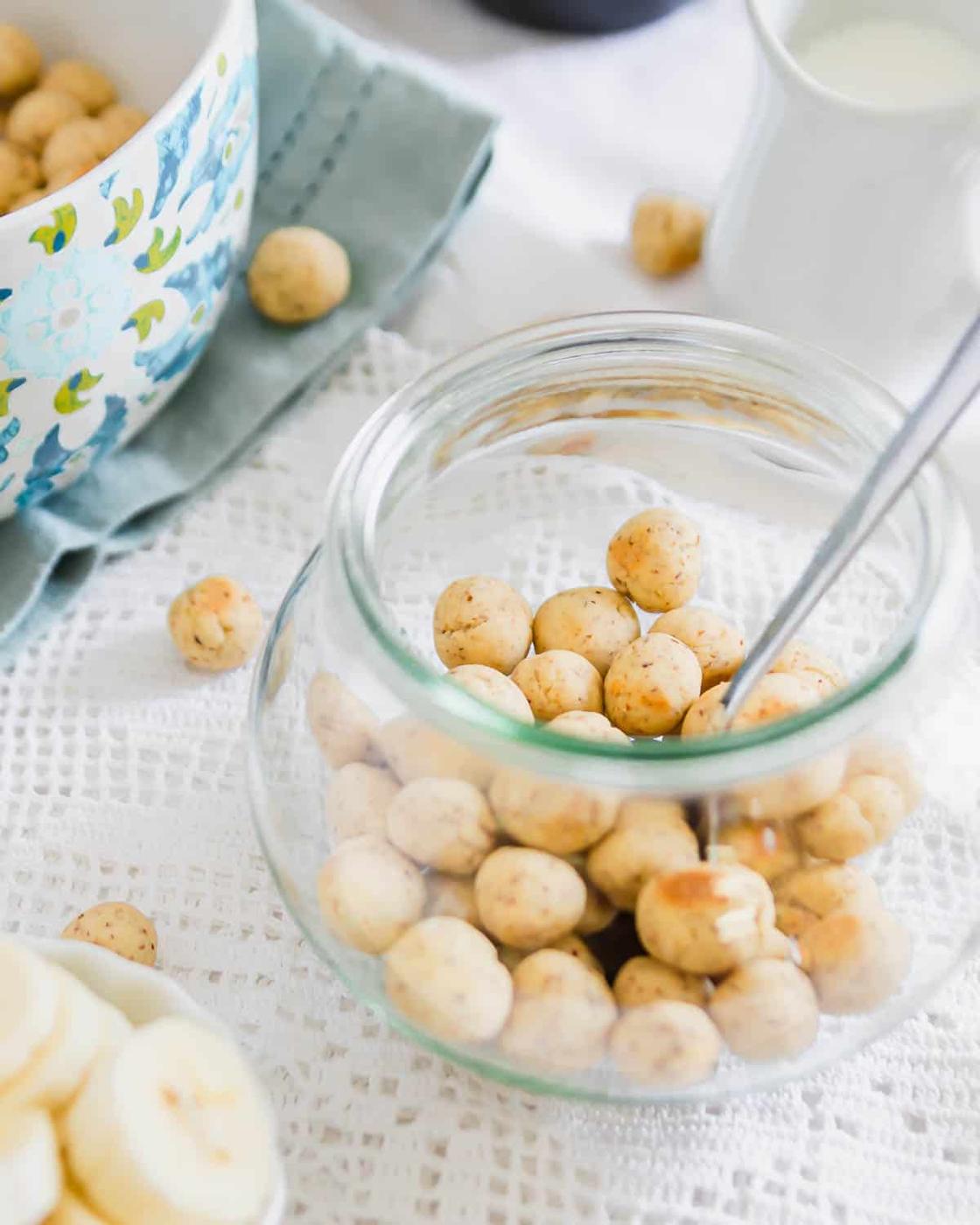 A jar filled with small, round cereal on a lace tablecloth. A spoon rests in the jar. Nearby, a bowl and a pitcher of milk are visible. A few snacks are scattered around the jar.