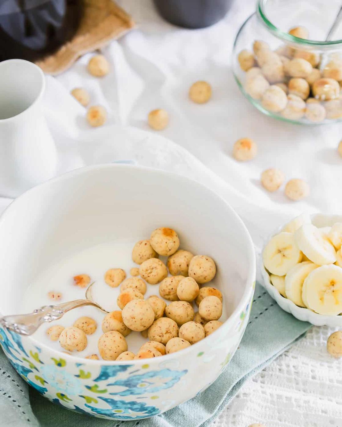 A bowl of milk with round cereal balls and a spoon on a light blue cloth. Sliced bananas in a small dish and more cereal scattered around. A glass jar and a jug are in the background on a white tablecloth.