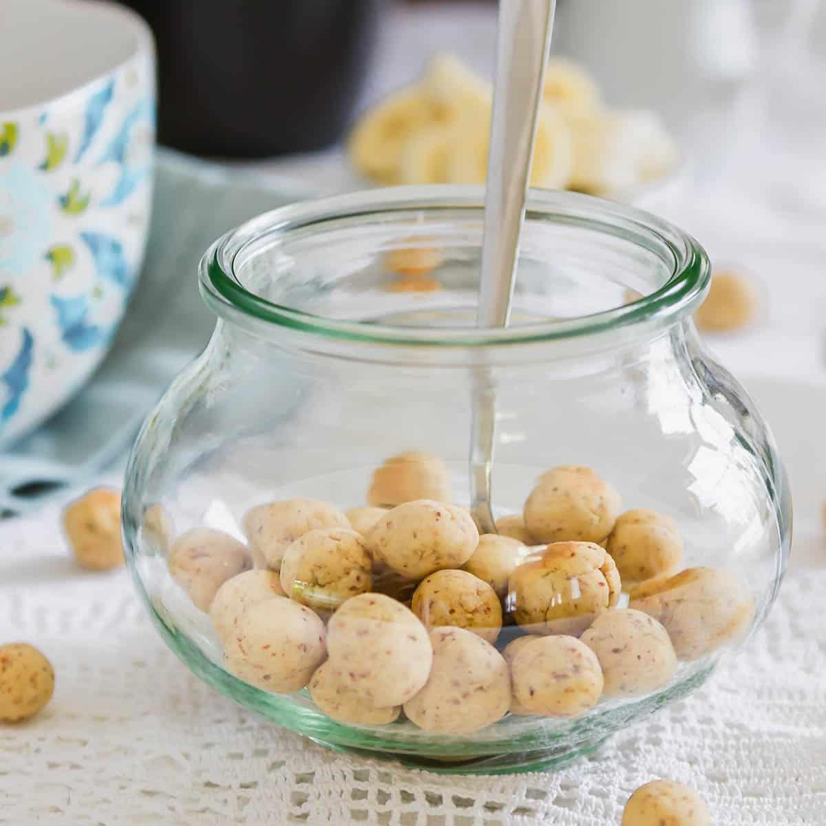 A glass jar filled with small round cereal balls. A spoon is inserted into the jar. A floral-patterned mug and sliced bananas are visible in the blurred background on a lace tablecloth.