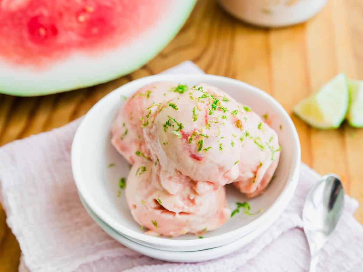 Three scoops of watermelon-flavored ice cream garnished with lime zest in a white bowl, set on a light cloth with a spoon. Watermelon and lime wedges are visible in the background.