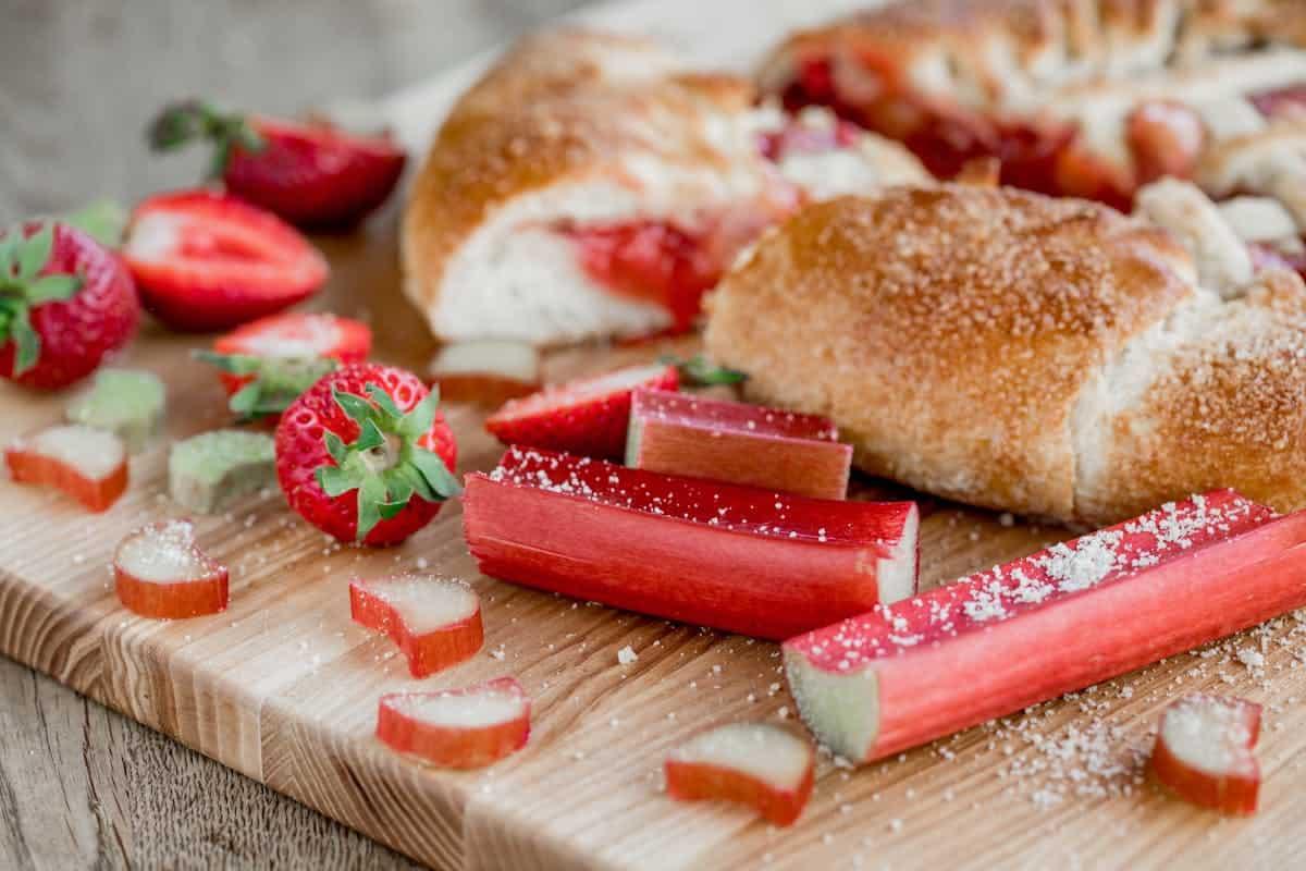 Sliced strawberries, rhubarb stalks (what is rhubarb?), and sugar-dusted pastries are displayed on a wooden cutting board.
